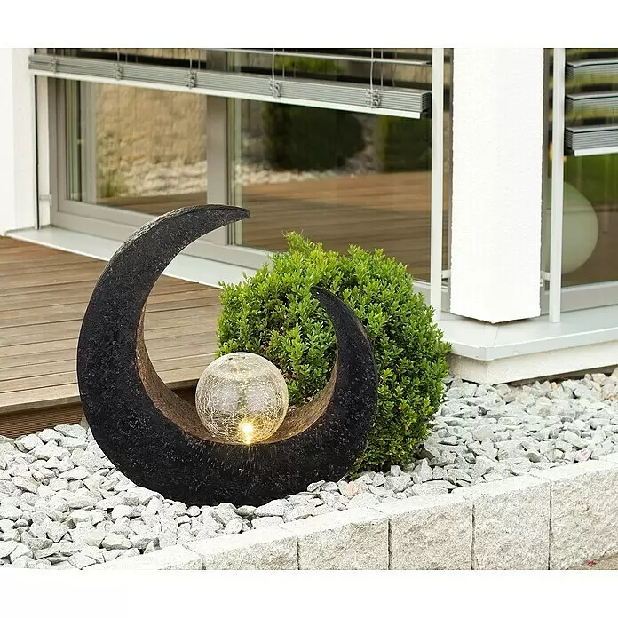 Globo Lámpara decorativa solar - 0,06 W, L x An x Al: 29,4 x 9 x 30,5 cm, Marrón, IP44 - Lámpara de jardín en forma de luna creciente gris oscuro con una esfera de vidrio craquelado brillante, entre grava y vegetación.