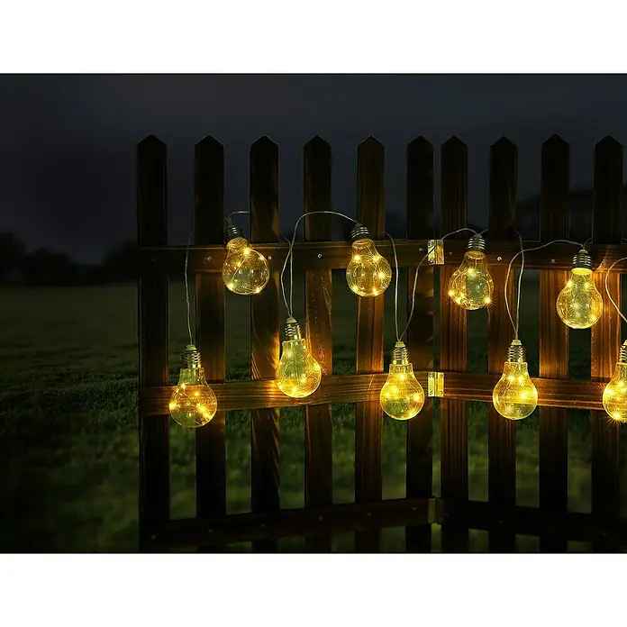 Globo Solar-Lichterkette - Klar - Bild einer Lichterkette mit Glühbirnen an einem Holzzaun im Garten, die eine warme Atmosphäre erzeugen.