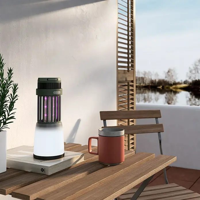 Coati Farolillo antimosquitos y luz portátil - 5 W, 40 m², Protección contra: Mosquitos - La imagen muestra una trampa para insectos en una mesa al aire libre, junto a una taza y un libro, con vistas a un lago.