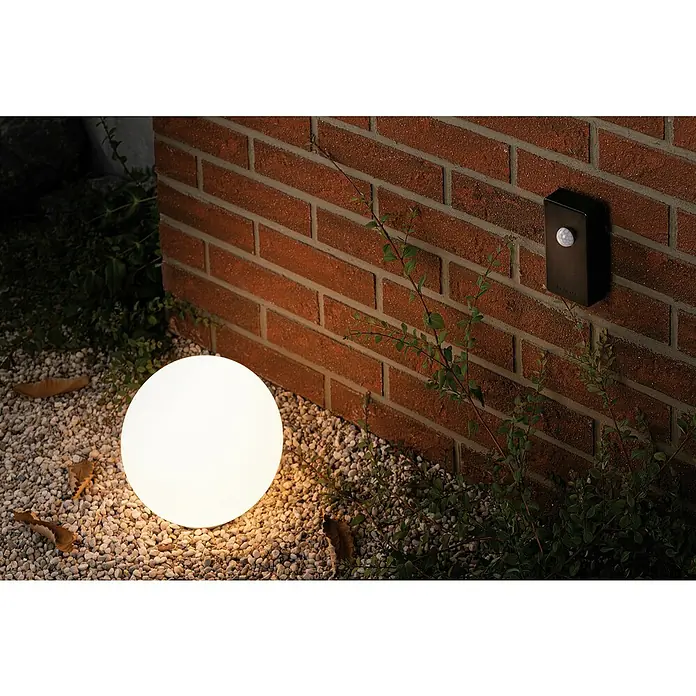 Paulmann Plug & Shine Sensor de movimiento - Antracita - Un foco esférico de exterior iluminado colocado sobre grava, iluminando una pared exterior, realzando el ambiente exterior.