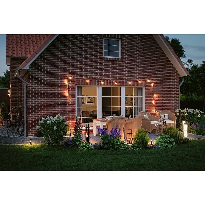 Paulmann Plug & Shine LED-Gartenstrahler Minipoller - Ø x H: 8 x 22 cm, 2 W, Anthrazit, IP44 - Die dekorative Lichterkette beleuchtet eine Backsteinfassade mit Terrassenmöbeln und üppiger Bepflanzung im Garten bei Dämmerung.