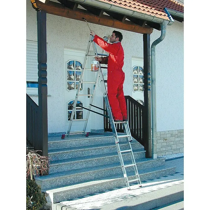 Krause Corda Mehrzweckleiter - Anzahl Tritte: 24 Stk., Arbeitshöhe: 5,4 m, Belastbarkeit: 150 kg, Aluminium, Treppengängig - Maler auf einer Klappleiter, der das Holz unter dem Dach streicht. Die Leiter steht auf einer Treppe vor dem Haus.