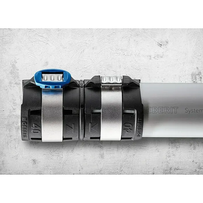 Geberit FlowFit Press-Übergangsstück auf Mepla 620.090.00.1 - Durchmesser 1: 16mm (Mepla), Durchmesser 2: 16mm (FlowFit) - Nahaufnahme einer Geberit Rohrverbindung mit blauer Verriegelung an einem grauen Rohr, befestigt an einer weißen Wand.