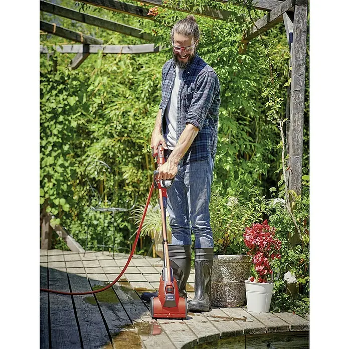 Einhell Power X-Change 18V Akku-Oberflächenbürste Picobella 18/215 - 18 V, Ohne Akku - Mann reinigt eine Holzterrasse mit einem elektrischen Terrassenreiniger, der mit einer Bürste und einem roten Schlauch ausgestattet ist.
