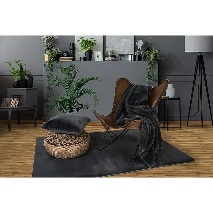 Decke HappyGraphit, 200 x 150 cm, 100 % Polyester - Ein grauer Teppich liegt unter einem braunen Ledersessel mit einer grauen Decke. Auf dem Teppich befindet sich auch ein Pouf mit einem Kissen.