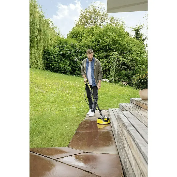 Kärcher Flächenreiniger T5 T-RacerPassend für: Kärcher Hochdruckreiniger K2 - K7 - Ein Mann benutzt einen Kärcher Flächenreiniger, um eine Terrasse zu reinigen. Der Flächenreiniger ist gelb und schwarz.