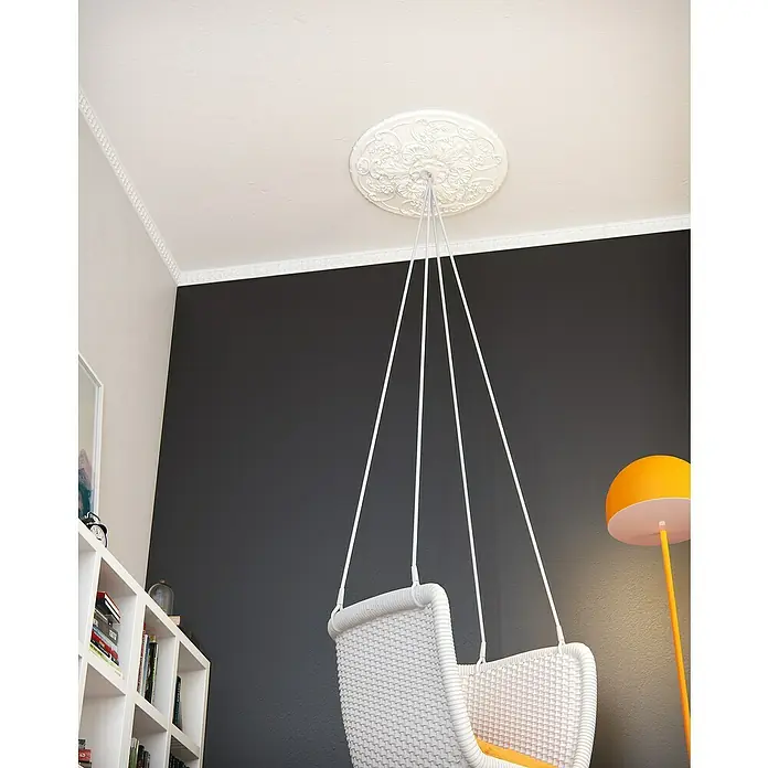 Nmc Decoflair Zierprofil Deckenleiste E21 - 2 m x 30 mm x 65 mm, Expandiertes Polystyrol (EPS) - Diese Aufnahme zeigt einen weißen Schaukelstuhl mit geflochtenem Design, der an einer dekorativen Deckenrosette hängt. Die helle Farbe des Stuhls steht im Kontrast zur dunklen Wand.