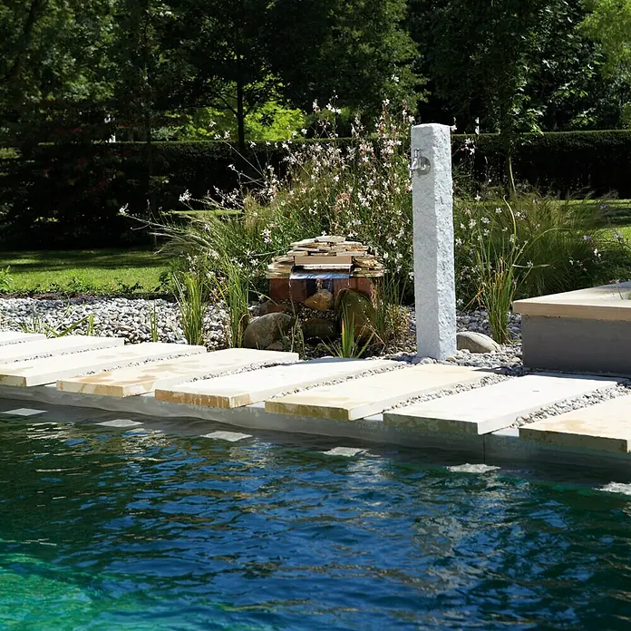 Garantia Wasserzapfstelle Granit - Lightgranite, Höhe: 100 cm - Gartenbrunnen mit Wasserhahn und Becken mit Steinplattenrand. Der Garten ist begrünt mit Bäumen, Sträuchern und Blumen.