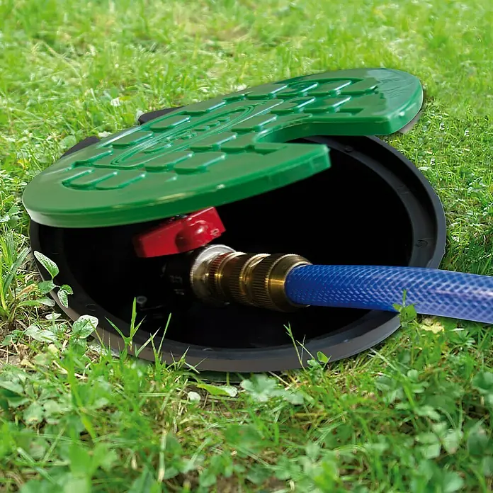 Arqueta redonda con tapa - Dimensión conexión: ¾″ - Una toma de agua de jardín subterránea con una manguera azul conectada a la conexión dorada y una tapa verde parcialmente abierta.