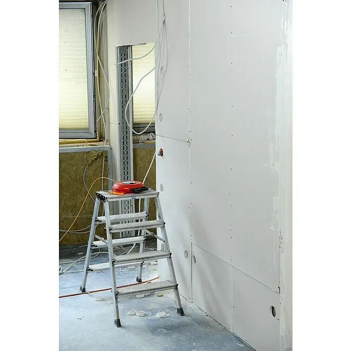 Krause Monto Doppel-Klapptritt Treppo - Anzahl Tritte: 8 Stk., Arbeitshöhe: 2,85 m, Aluminium - Eine silberne Leiter mit einem roten Gerät darauf steht in einem unfertigen Raum mit angebrachten Trockenbauwänden und freiliegenden Kabeln.
