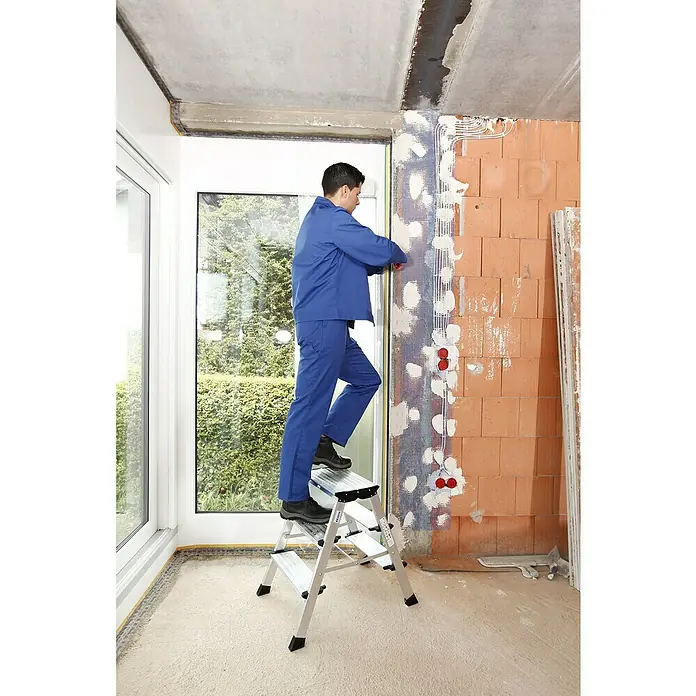 Krause Monto Doppel-Klapptritt Treppo - Anzahl Tritte: 6 Stk., Arbeitshöhe: 2,65 m, Aluminium - Ein Handwerker auf einer KRAUSE Leiter befestigt ein Netz an einer Ziegelwand in einem Raum mit einem großen Fenster.