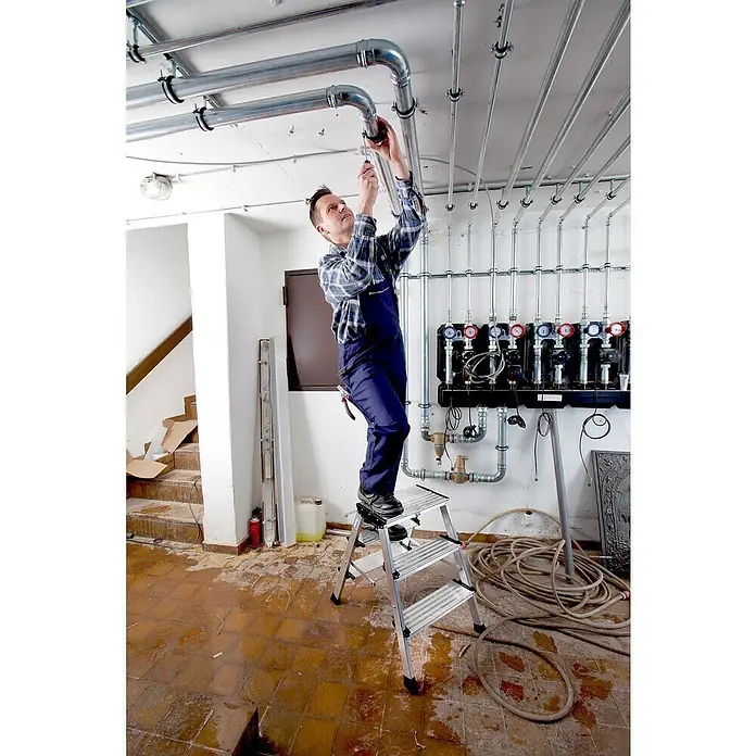 Krause Monto Doppel-Klapptritt Treppo - Anzahl Tritte: 6 Stk., Arbeitshöhe: 2,65 m, Aluminium - Ein Mann auf einer silbernen Trittleiter montiert Rohre an einer Decke. Er trägt blaue Arbeitskleidung und ein kariertes Hemd.