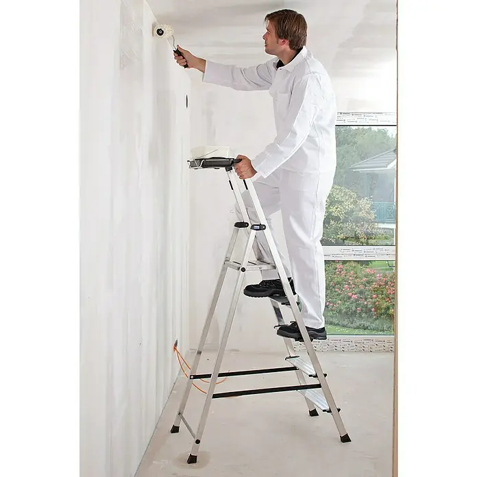 Krause Monto Stehleiter Secury MultiGrip - Anzahl Tritte: 5 Stk., Arbeitshöhe: 3,05 m, Belastbarkeit: 150 kg, Aluminium - Ein Mann im weißen Anzug steht auf einer silbernen Leiter und streicht eine Wand mit einer Farbrolle weiß.