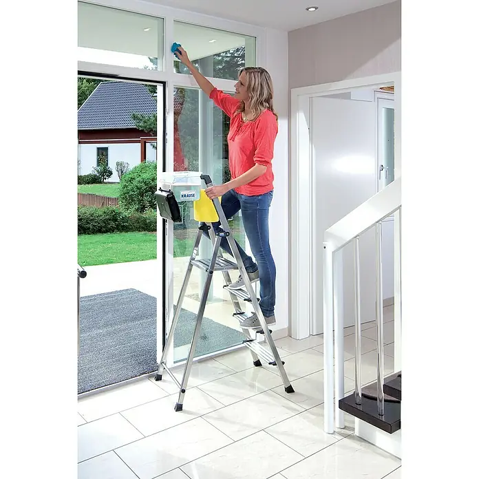 Krause Monto Stehleiter Secury MultiGrip - Anzahl Tritte: 4 Stk., Arbeitshöhe: 2,85 m, Belastbarkeit: 150 kg, Aluminium - Frau putzt Fenster stehend auf einer Krause Tritthocker Leiter. Die Leiter ist leicht und einfach zu verstauen.