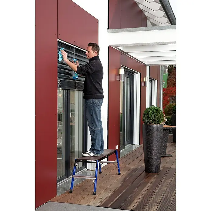 Krause Monto Arbeitsplattform StepTop - Anzahl Tritte: 1 Stk., Arbeitshöhe: 2,45 m, Aluminium - Ein Mann reinigt Rollläden von einem Tritt aus. Die Trittstufenleiter ist blau und schwarz. Im Hintergrund befindet sich eine Terrasse.