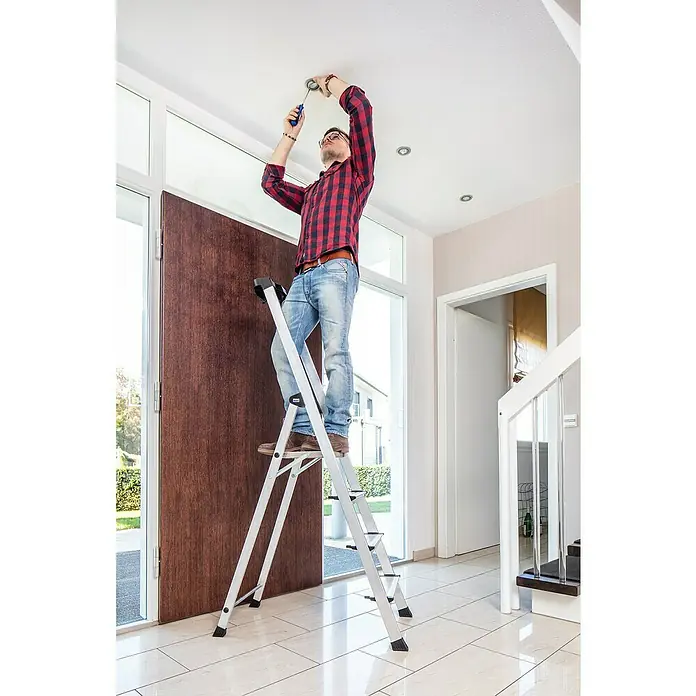 Krause PlusLine Stehleiter Safety - Anzahl Tritte: 4 Stk., Arbeitshöhe: 2,85 m, Aluminium - Ein Mann steht auf einer Aluminium-Haushaltsleiter und repariert mit einem Schraubenzieher eine Deckenleuchte in einem modernen Haus.