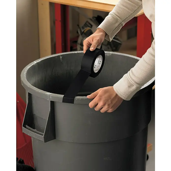 3M Cinta adhesiva universal Scotch americana - Negro, An x L: 48 mm x 25 m, 1 ud. - Una persona pega cinta adhesiva negra a un bote de basura gris, posiblemente para hacer una reparación o asegurar algo.