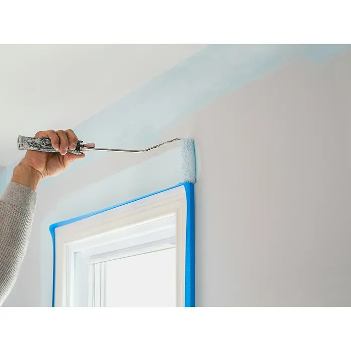 3M Cinta de enmascarar ScotchBlue - Azul, An x L: 24 mm x 41 m, 1 ud. - Un trabajador pinta una franja azul claro en una pared blanca con un rodillo de pintura. Las ventanas están protegidas con cinta azul.