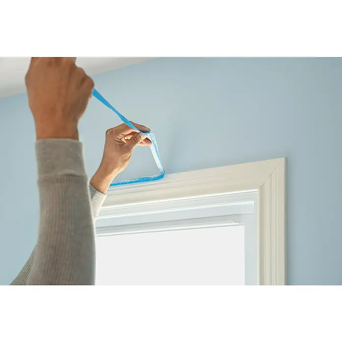 3M Cinta de enmascarar ScotchBlue - Azul, An x L: 24 mm x 41 m, 1 ud. - Una persona aplica cinta de pintor azul a un marco de ventana blanco para proteger la pared durante la pintura.