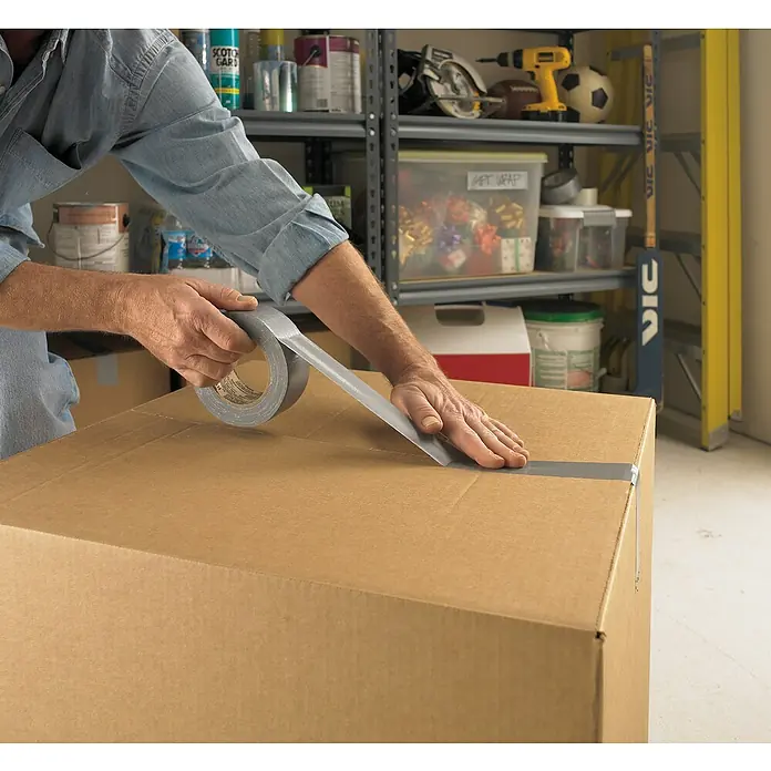 3M Cinta adhesiva universal Scotch americana - Gris, An x L: 48 mm x 25 m, 1 ud. - Primer plano de una persona sellando una caja de cartón con cinta adhesiva gris, con una estantería llena de herramientas al fondo.