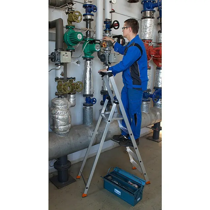 Krause Monto Stehleiter Solido - Anzahl Tritte: 5 Stk., Arbeitshöhe: 3,05 m, Aluminium - Ein Handwerker steht auf einer Krause-Leiter vor einer Wand mit Heizungsrohren und Armaturen, um Wartungsarbeiten durchzuführen.