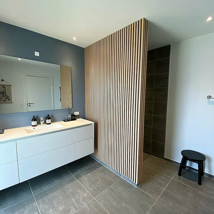 FibroTech Akustikpaneel Unika 25 - Light Oak, 2.440 x 600 x 22 mm - Badezimmer mit modernem Waschtisch, großem Spiegel und einer stilvollen Trennwand aus Holzlamellen für eine elegante Raumaufteilung.