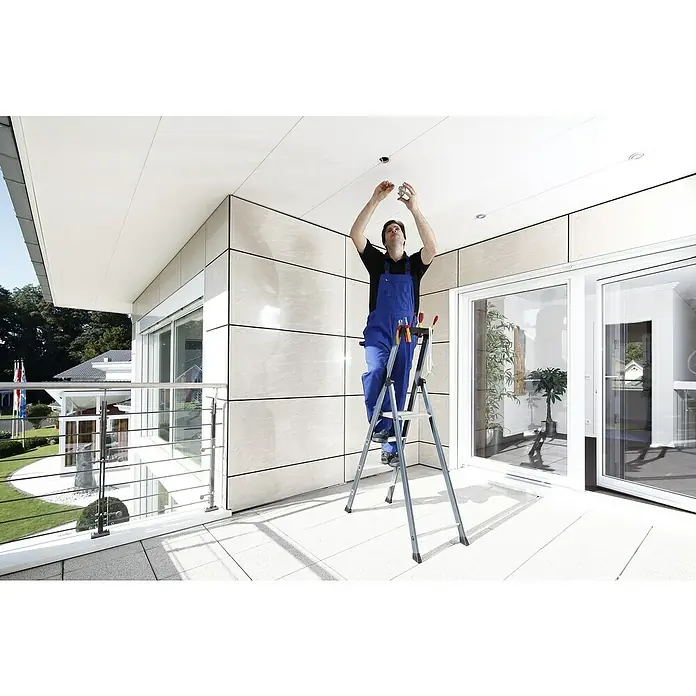 Krause Monto Stehleiter SePro S - Anzahl Tritte: 4 Stk., Arbeitshöhe: 2,85 m, Aluminium - Ein Handwerker auf einer Leiter installiert eine Deckenleuchte, wobei er blaue Arbeitskleidung trägt und Werkzeuge bei sich hat.