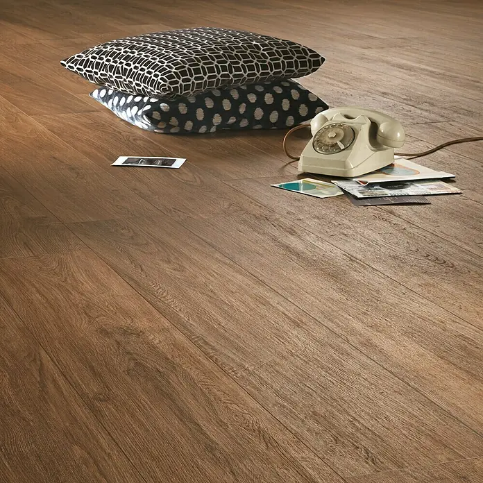Harmonie by Palazzo Feinsteinzeugfliese - 30 x 120 cm, Eiche Dunkel, Matt - Brauner Laminatboden mit Kissen, Telefon und Fotos. Die Laminatstruktur ahmt echtes Holz nach und sorgt für eine warme, einladende Atmosphäre.