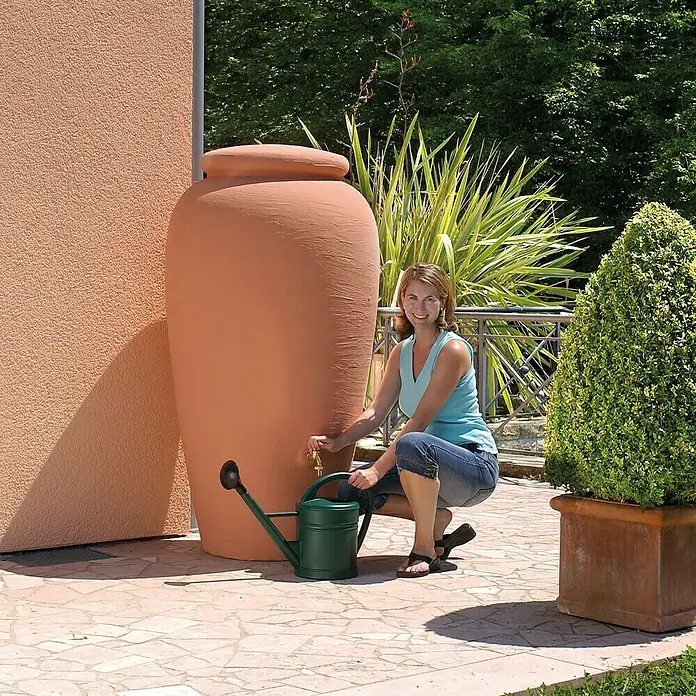 Garantia Classic Line Regenspeicher Amphore - 500 l, Terrakotta - Eine Frau füllt eine grüne Gießkanne aus einem Terrakotta-Regenwassertank im Freien. Der Terrakotta-Tank ist ideal für die Wasserspeicherung.