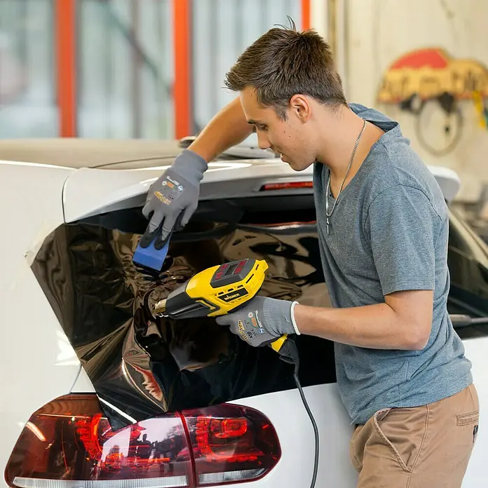 Wagner Heißluftgebläse Furno 750 - 2.000 W - Ein Mann verwendet eine Wagner Heißluftpistole, um eine dunkle Folie auf einem weißen Auto anzubringen, wobei er Handschuhe und einen Rakel benutzt.