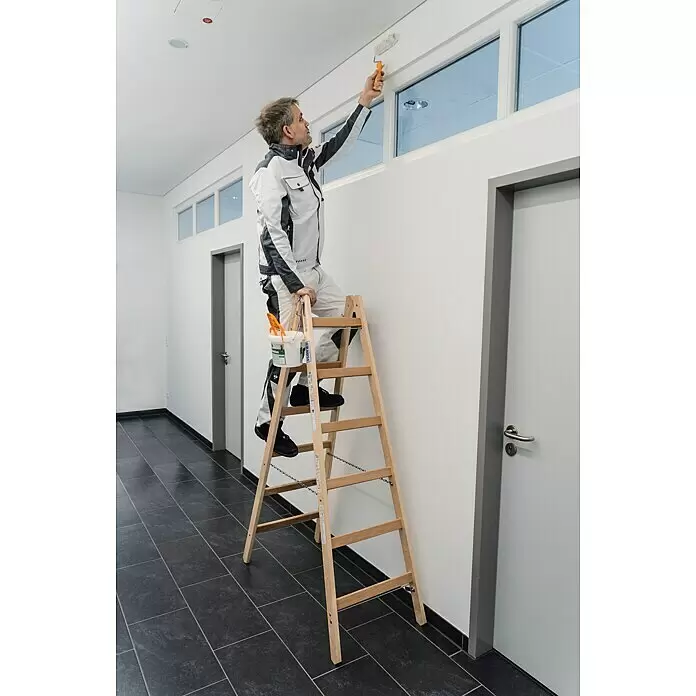 Krause Stabilo Escalera doble - Número de peldaños: 12 ud., Altura de trabajo: 3 m, Madera - Escalera de tijera de madera en uso. Un pintor está de pie sobre ella, alcanzando un marco de ventana con un rodillo.