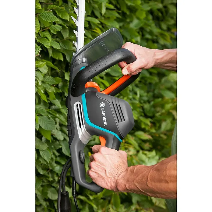 Gardena PowerCut Elektro-Heckenschere 700/65 - 700 W, Schnittlänge: 65 cm - Eine Gardena Heckenschere wird von zwei Händen vor einer Hecke gehalten, die Heckenschere ist grau, schwarz und orange.