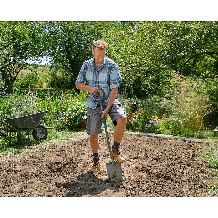 Gardena Ergoline Gärtnerspaten - Ergonomischer Stiel, Länge Stiel: 106 cm - 126 cm - Ein Mann benutzt einen Gardena Spaten in einem Garten, um den Boden vorzubereiten, mit einem Schubkarren und grüner Vegetation im Hintergrund.