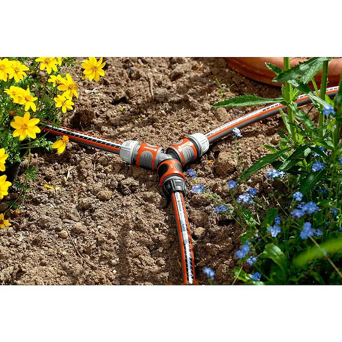 Gardena Y-Stück - Anschlüsse: 3 - Ein Gardena-Verteilerstück, das ein Bewässerungssystem mit Gartenschläuchen auf einem Erdboden mit Pflanzen und Blumen verbindet.