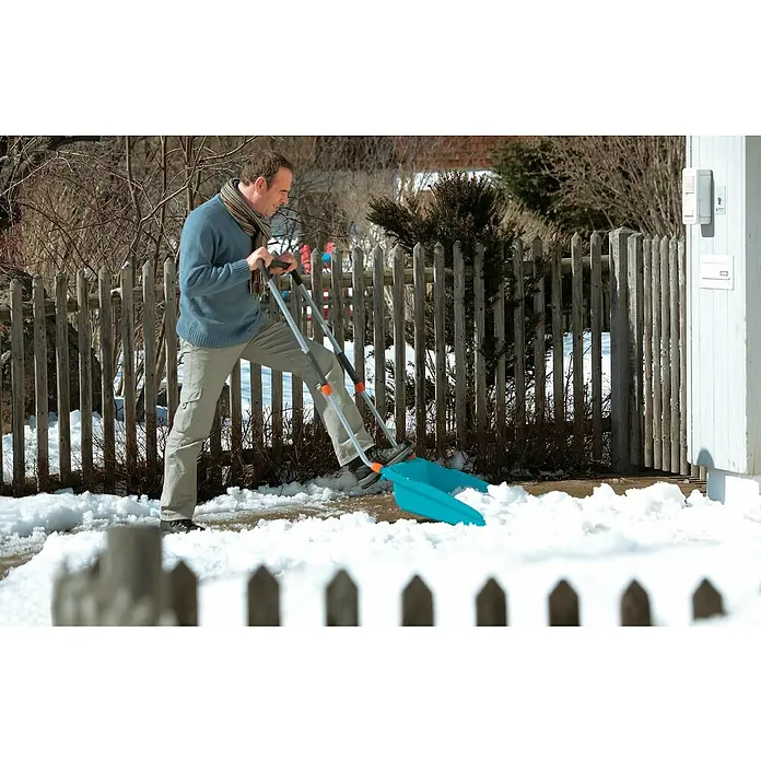 Gardena Schneewanne - Breite: 70 cm, Teleskopstiel, Kunststoff - Ein Mann benutzt eine blaue Schneeschaufel mit Rädern, um Schnee vor einem Haus mit weißem Zaun zu räumen.