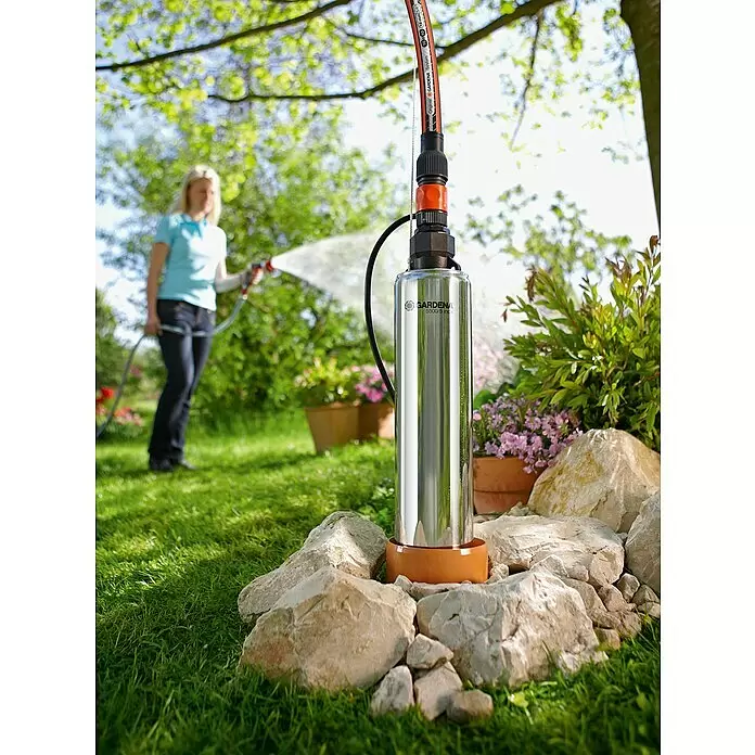 Gardena Tiefbrunnenpumpe 5500/5 Inox - 850 W, 5.500 l/h, Edelstahl - Edelstahl-GARDENA-Pumpe mit Schlauch, im Garten stehend, während eine Frau im Hintergrund Pflanzen bewässert.