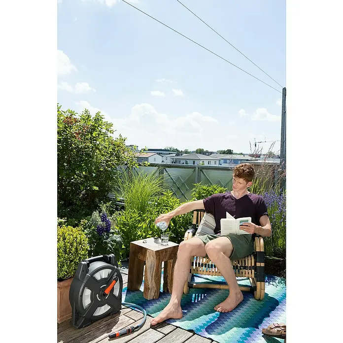 Gardena Schlauchtrommel - Schlauchlänge: 10 m, Kunststoff - Ein Mann sitzt im Garten und liest ein Buch, während er Wasser in ein Glas gießt. Im Vordergrund ist eine GARDENA Schlauchbox zu sehen.