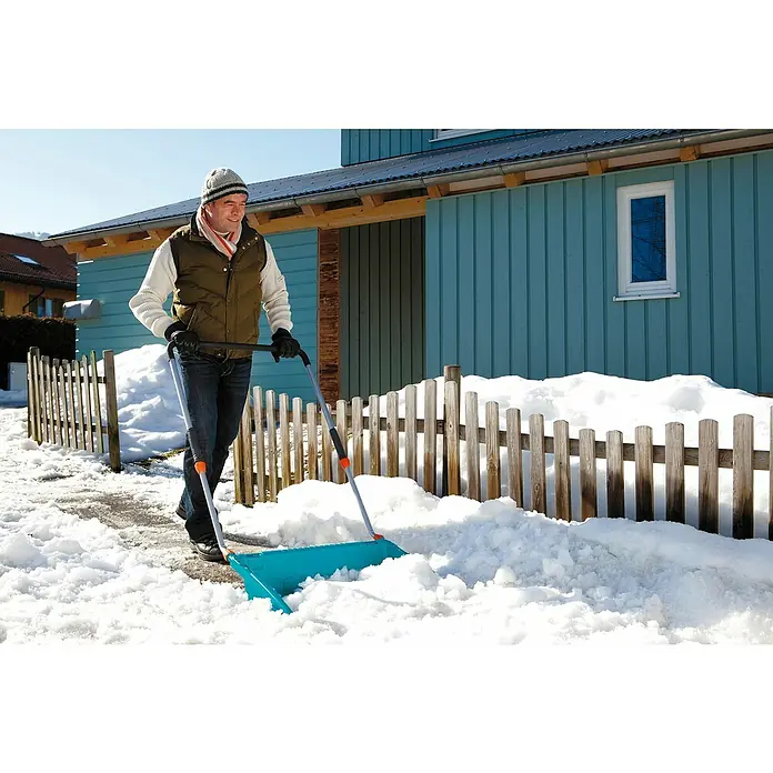 Gardena Schneewanne - Breite: 70 cm, Teleskopstiel, Kunststoff - Mann schiebt mit einem blauen Schneeschieber Schnee vor einem hellblau gestrichenen Haus und einem Holzzaun.