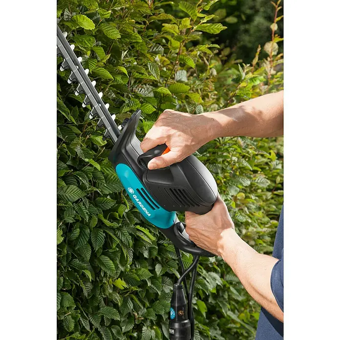 Gardena EasyCut Elektro-Heckenschere 500/55 - 500 W, Schnittlänge: 55 cm, Max. Aststärke: 18 mm - Nahaufnahme einer Person, die eine Gardena-Heckenschere an einer grünen Hecke benutzt, um die Äste zu schneiden und die Form zu erhalten.