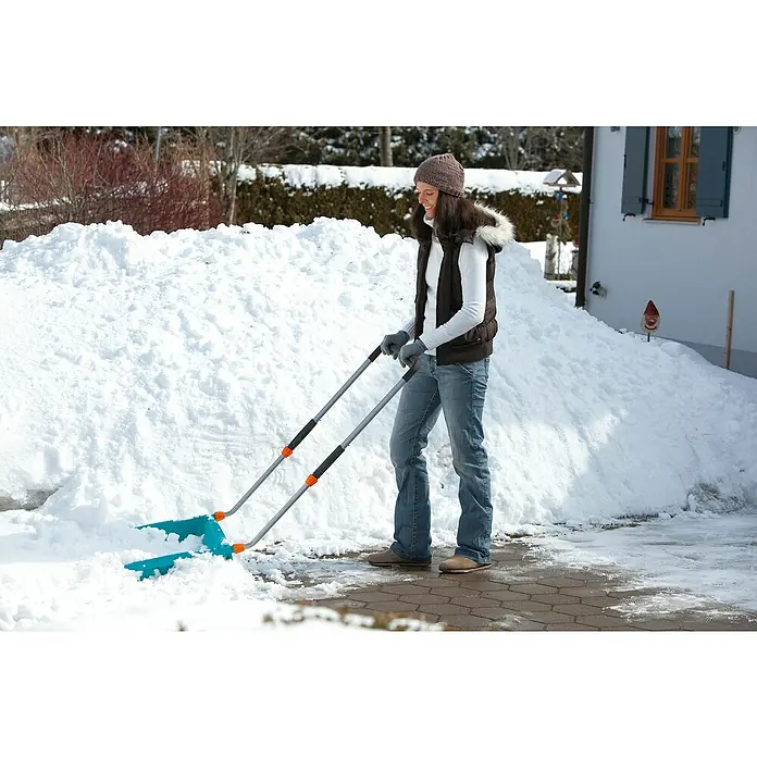 Gardena Schneewanne - Breite: 70 cm, Teleskopstiel, Kunststoff - Frau räumt mit einer blauen Schneeschaufel Schnee von einer gepflasterten Auffahrt. Sie trägt Mütze, Weste, Jeans und Handschuhe.