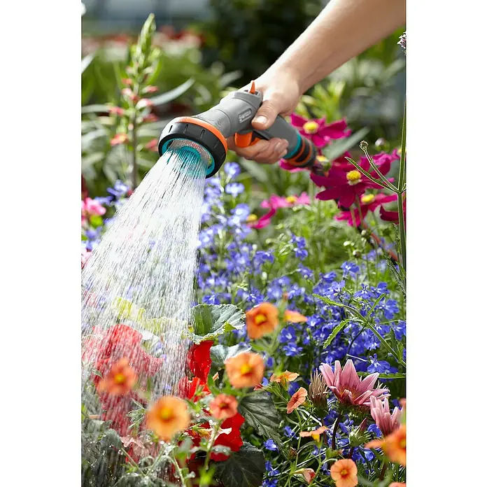 Gardena Comfort Vielflächenregner - Anzahl Sprühformen: 2 Stk., Kunststoff - Nahaufnahme einer Hand, die mit einer Gardena-Gartenspritze bunte Blumen bewässert, die in voller Blüte stehen, während Wassertropfen in der Luft hängen.