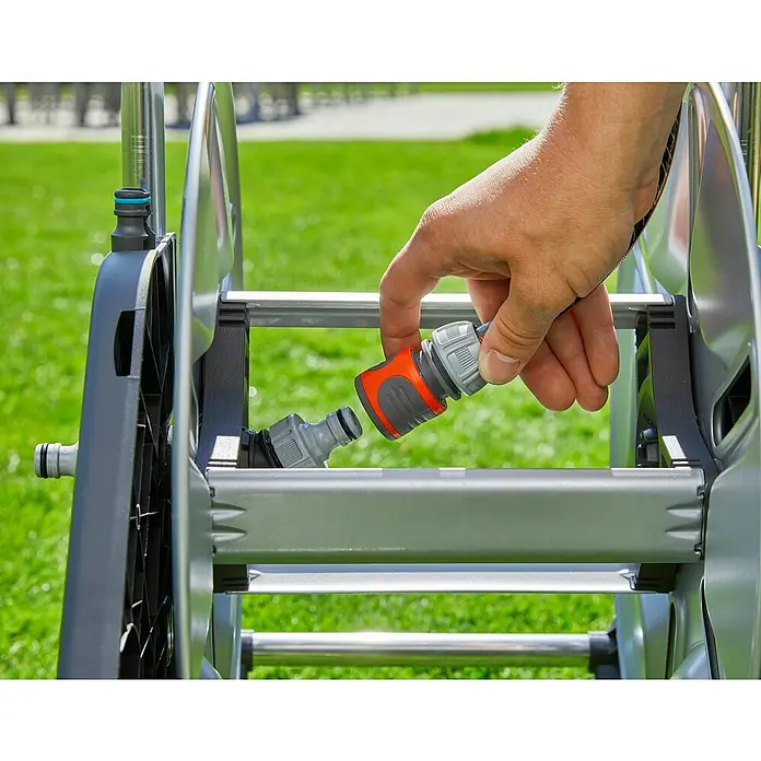 Gardena Schlauchwagen-Set CleverRoll M - Kunststoff/Metall, Schlauchlänge: 25 m - Ein Mann verbindet ein orangefarbenes Gartenschlauchstück mit einem grauen Teil eines Schlauchaufrollers im Freien auf Rasen.
