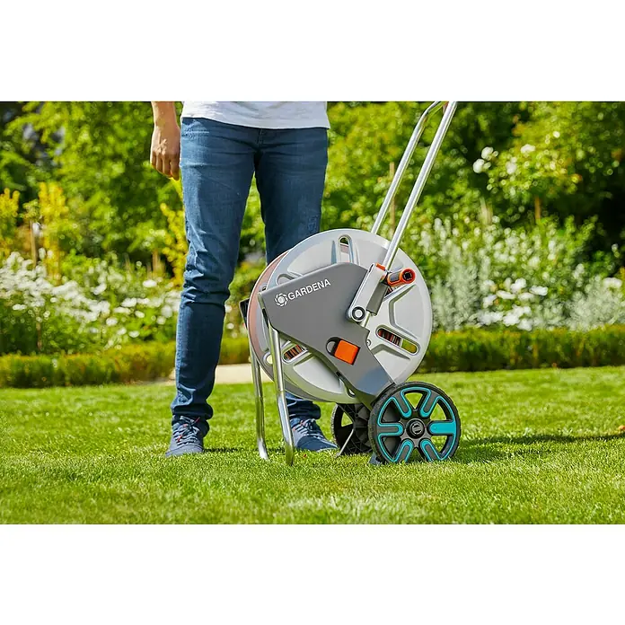 Gardena Schlauchwagen CleverRoll L Easy - Metall, Schlauchkapazität: 100 m, 13 mm (½″) - Eine Person steht neben einer GARDENA Gartenschlauchbox mit Rädern auf einem grünen Rasen.