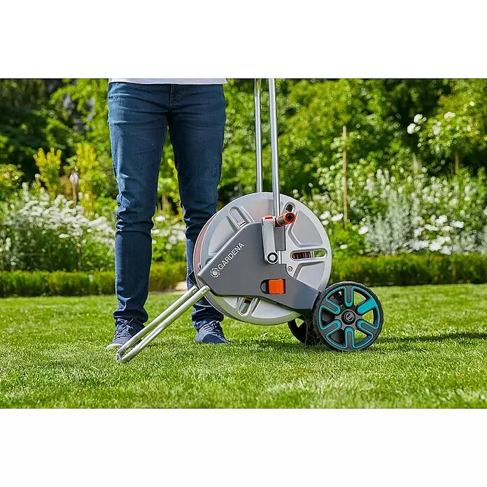 Gardena Schlauchwagen-Set CleverRoll M - Kunststoff/Metall, Schlauchlänge: 25 m - Grauer und oranger Gardena Schlauchwagen mit Rädern und Standfuß, in einem Garten mit einer Person.