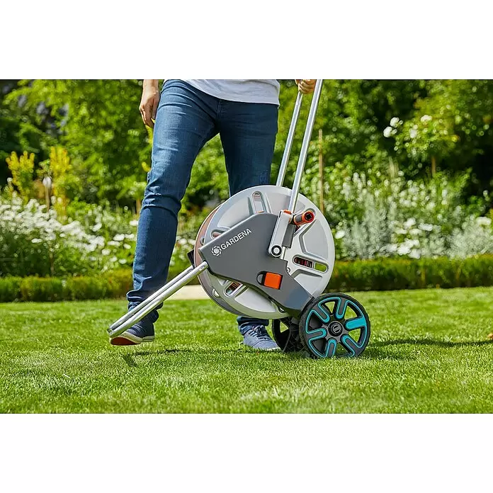 Gardena Schlauchwagen CleverRoll L Easy - Metall, Schlauchkapazität: 100 m, 13 mm (½″) - Eine Person schiebt eine grau-silberne Gardena Schlauchtrommel mit schwarz-türkisen Rädern auf einem Rasen.