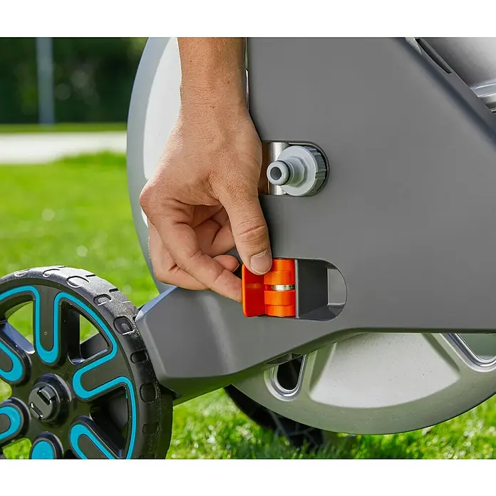 Gardena Carro portamangueras Aquaroll L Easy - Metal, Capacidad de manguera: 100 m, 13 mm (½'') - Primer plano de una mano que fija una pieza naranja a un dispositivo de jardinería Gardena, situado sobre un césped.