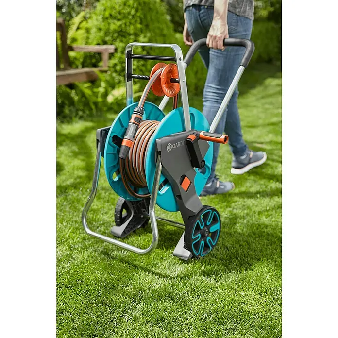 Gardena Schlauchwagen-Set CleverRoll M Easy Set - Kunststoff/Metall, Schlauchlänge: 20 m - Ein Rasen mit einem Gardena-Schlauchwagen mit Schlauchaufroller, der von einer Person gezogen wird. Der Wagen ist blau, grau und orange.