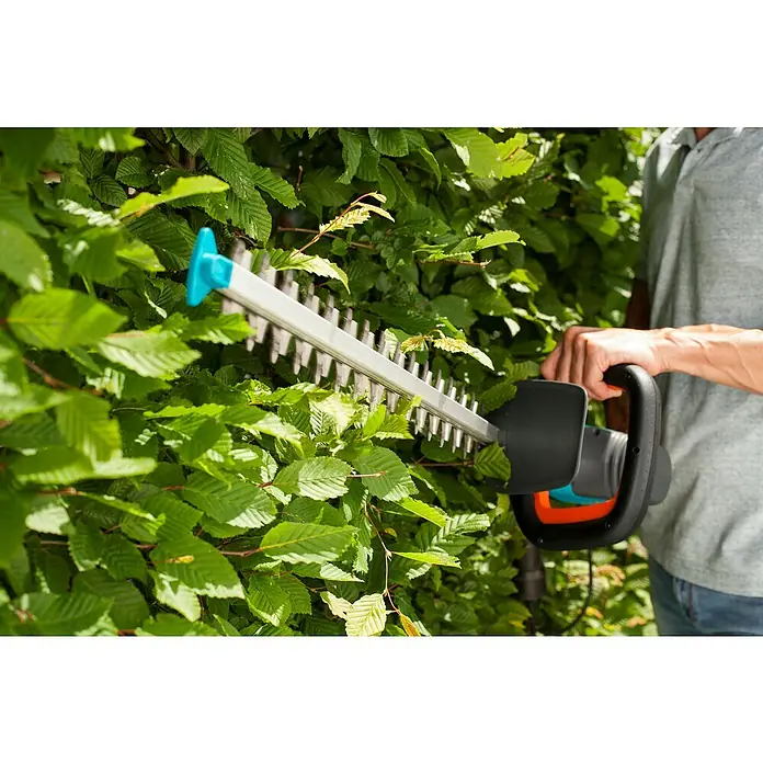 Gardena ComfortCut Elektro-Heckenschere 600/55 - 600 W, Schnittlänge: 55 cm - Eine Heckenschere von Gardena schneidet eine grüne Hecke. Das Gerät ist blau, grau und orange und wird von einer Person bedient.