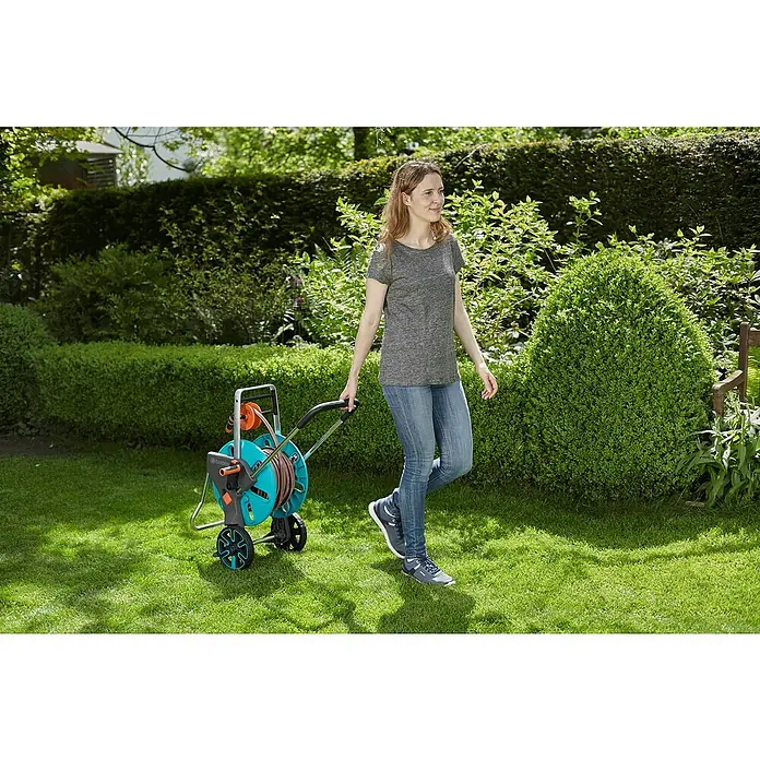Gardena Schlauchwagen-Set CleverRoll M Easy Set - Kunststoff/Metall, Schlauchlänge: 20 m - Eine Frau zieht einen Gardena Schlauchwagen durch einen grünen Garten. Der Schlauchwagen ist mit einem orangefarbenen Schlauch und Rädern versehen.