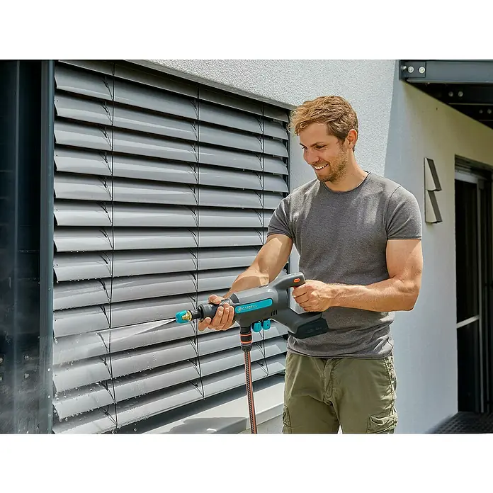 Gardena Power for All 18V Akku-Druckreiniger AquaClean 24/18V SE - 18 V, 1 Akku, 2,5 Ah - Ein Mann reinigt Fensterläden mit dem Gardena Akku-Reiniger. Das Gerät ist blau-grau und wird mit einem Akku betrieben. Es sprüht Wasser auf die Lamellen.